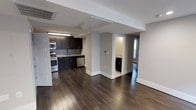 This single-floor space showcases a thoughtfully designed open-concept layout that seamlessly connects the main living area to the kitchen. Rich, dark hardwood flooring extends throughout, providing a warm and cohesive aesthetic that contrasts elegantly with the crisp white walls and baseboards.

The kitchen is well-appointed with dark espresso cabinetry that reaches toward the ceiling, offering generous storage. A gray tile backsplash adds texture behind the countertops, while stainless steel appliances — including a gas range, microwave, and dishwasher — provide a modern, functional edge. A utility sink anchors the workspace, completing the kitchen's practical design.

A structural column and partial wall divide the open space, creating a natural visual separation between the kitchen and living areas without compromising the open feel. Beyond the column, a hallway leads to additional rooms, including what appears to be a bathroom with classic black-and-white tile flooring and a separate room benefiting from natural light through a window. Recessed and flush-mount lighting fixtures ensure the space remains well-lit throughout.