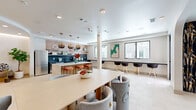 Located at 701 Richmond Ave in Houston's Montrose neighborhood, this single-floor amenity space offers a well-appointed communal environment thoughtfully divided into distinct functional zones. The open-plan layout accommodates a lounge area, a dining/gathering table, a bar-style counter with high stools, and a fully equipped kitchenette—all unified by light-toned large-format tile flooring that runs throughout.

The kitchenette is a standout feature, outfitted with dark matte cabinetry, a concrete-style countertop with an undermount sink, stainless steel appliances including a full-size refrigerator, and a striking two-tone tile backsplash combining charcoal subway tiles below and warm rose-toned square tiles above. Open floating shelves add both storage and decorative display space. A terrazzo island with pendant lighting overhead anchors the transition between the kitchen and dining areas.

The lounge zone is anchored by a bold patterned feature wall hosting a wall-mounted flat-screen TV, complemented by an ornate sculptural chandelier. Built-in open shelving displays books and décor, while mid-century-inspired seating and layered rugs create a comfortable, curated atmosphere. Large windows along one wall allow natural light to filter in, and floor-to-ceiling curtains frame views to an outdoor terrace, enhancing the sense of openness throughout the space.