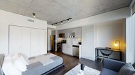 This single-floor studio showcases a striking industrial-modern design, defined by its exposed concrete ceiling that lends an urban, loft-like character to the space. Dark hardwood flooring runs throughout, providing a warm contrast to the cool concrete overhead and the crisp white walls and cabinetry.

The open-plan layout seamlessly integrates the sleeping and living areas with a well-appointed kitchen. The kitchen features sleek, flat-front white cabinetry, stainless steel appliances including a refrigerator, gas range, built-in microwave, and dishwasher, along with a clean white tile backsplash and a single-basin sink. The cabinetry extends to the ceiling, maximizing vertical storage.

A track lighting system mounted to the concrete ceiling provides adjustable, directional illumination throughout the space. The sleeping area accommodates a full-sized bed with a low-profile frame, complemented by a plush area rug. A compact desk and chair are tucked near one wall, offering a functional workspace. Built-in closet storage with floor-to-ceiling white panel doors contributes to the space's clean, uncluttered aesthetic.