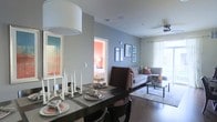 This well-appointed single-floor residence offers a thoughtfully designed open-concept layout that seamlessly connects the dining and living areas, creating a sense of spaciousness and flow throughout the home. The dining area features a dark-toned table set for entertaining, complemented by a striking piece of wall art that adds a vibrant pop of color to the space. Adjacent to the dining area, the living room is anchored by a comfortable sectional sofa accented with decorative pillows, a sleek glass coffee table, and a soft area rug that defines the seating area. Framed artwork adorns the walls, lending a curated, gallery-like quality to the space. A ceiling fan with an integrated light fixture overhead adds both function and style. Natural light filters through sheer curtains covering a large window, brightening the room and offering a view to the outdoors. The cool gray wall tones throughout create a calm, modern ambiance that ties the spaces together cohesively. A glimpse into the bedroom reveals a warm coral-toned accent wall and soft lighting, providing a cozy retreat. Rich hardwood-style flooring runs throughout, adding warmth and continuity to the overall design aesthetic.