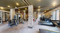Located at 215 Reformation Pkwy in Canton, GA, this expansive fitness center offers a generously sized, single-floor exercise space designed to support a wide range of workout routines. The room's architectural character is defined by striking exposed brick walls and structural brick columns that divide the floor plan into distinct workout zones while maintaining an open, airy feel. A dark coffered ceiling with wood beam detailing and recessed lighting adds a refined, industrial-chic aesthetic throughout.

The cardio zone is well-appointed with multiple treadmills, elliptical machines, and upright bikes, several of which feature built-in digital displays. Natural light filters in through several windows along the exterior walls, offering views of the surrounding greenery. Wall-mounted fans provide additional ventilation and comfort.

The strength training area is equally comprehensive, featuring a multi-station cable machine, selectorized weight machines, a full dumbbell rack, and a variety of free weights. Functional training accessories — including kettlebells, medicine balls, stability balls, and a dedicated storage rack — are neatly organized and readily accessible. A rowing machine and adjustable weight benches round out the available equipment. The durable, patterned flooring ties the space together, creating a functional and visually cohesive fitness environment.