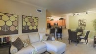 This single-floor residence offers an open-concept layout that seamlessly connects the living area to the kitchen, creating a spacious and cohesive environment. The living room is anchored by a plush sectional sofa adorned with decorative pillows, complemented by soft carpeting and neutral-toned walls accented with crown molding. A bold, nature-inspired wall art piece adds a striking focal point to the space. Track lighting overhead ensures the area is well-lit and inviting.

The kitchen is designed for both functionality and style, featuring warm honey-toned cabinetry, a dark granite or stone-finish breakfast bar with bar seating, and stainless steel appliances including a refrigerator and microwave. The raised bar counter serves as a natural divider between the kitchen and living areas while maintaining an open, airy feel throughout.

Wainscoting details and crisp white trim along the walls and baseboards add a polished, finished look to the interior. The overall color palette of soft greens, warm browns, and creamy neutrals creates a cohesive and calming ambiance. The thoughtful layout and design details make this a comfortable and practical single-floor living space.