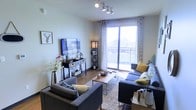 This single-floor residence offers a thoughtfully designed living space that balances comfort and contemporary style. The living room serves as the focal point of the home, featuring warm hardwood flooring, neutral-toned walls, and a modern track lighting system on the ceiling that provides flexible, even illumination throughout the room. Large sliding glass doors flood the space with abundant natural light and open directly onto a private balcony, offering open views of the surrounding cityscape. The living area is generously proportioned, comfortably accommodating a sectional sofa arrangement centered around a sleek coffee table and area rug. A wall-mounted flat-screen television is paired with a media console, creating a well-organized entertainment setup. Decorative accents, including floral arrangements, accent pillows in muted gold tones, and tasteful wall art, contribute to the warm and inviting ambiance. Patterned curtains frame the balcony doors, adding a layer of texture and visual interest. The overall atmosphere is modern, airy, and welcoming, with a cohesive design that makes excellent use of the available space while maximizing natural light.
