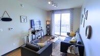 This single-floor residence offers a thoughtfully designed living space that balances comfort and contemporary style. The living room serves as the focal point of the home, featuring warm hardwood flooring, neutral-toned walls, and a modern track lighting system on the ceiling that provides flexible, even illumination throughout the room. Large sliding glass doors flood the space with abundant natural light and open directly onto a private balcony, offering open views of the surrounding cityscape. The living area is generously proportioned, comfortably accommodating a sectional sofa arrangement centered around a sleek coffee table and area rug. A wall-mounted flat-screen television is paired with a media console, creating a well-organized entertainment setup. Decorative accents, including floral arrangements, accent pillows in muted gold tones, and tasteful wall art, contribute to the warm and inviting ambiance. Patterned curtains frame the balcony doors, adding a layer of texture and visual interest. The overall atmosphere is modern, airy, and welcoming, with a cohesive design that makes excellent use of the available space while maximizing natural light.