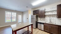 This efficient single-floor, one-bedroom, one-bathroom unit is thoughtfully designed to maximize functionality and comfort. The open-concept kitchen serves as the heart of the space, showcasing rich dark espresso cabinetry, granite countertops, and a full suite of stainless steel appliances including a refrigerator, dishwasher, microwave, and range. A decorative mosaic tile backsplash adds a stylish accent, while a freestanding granite-topped island provides additional prep and dining space. Light hardwood-style flooring runs throughout the kitchen and living areas, creating a warm and cohesive aesthetic. Natural light filters in through windows near the entryway, enhancing the open feel of the main living space. The bedroom is a comfortable retreat, featuring neutral carpet flooring, soft gray walls, and a generously sized window that brings in ample natural light and outdoor views. A dedicated closet offers practical storage. The bathroom is clean and well-finished, with a granite-topped dark vanity, a frameless mirror, a tub-shower combination, and matching hardwood-style flooring that ties the space together. The overall design is modern and consistent, with neutral tones and quality finishes throughout.