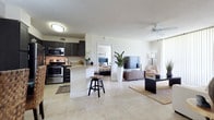 This single-floor residence offers a thoughtfully designed open-concept layout that seamlessly connects the kitchen, dining, and living areas, creating a bright and cohesive living environment. The kitchen is equipped with dark cabinetry, stainless steel appliances including a refrigerator, electric range, and microwave, complemented by granite-style countertops and a peninsula with bar seating — ideal for casual dining or entertaining. Adjacent to the kitchen, the dedicated dining area accommodates a full table set beneath a pendant light fixture, with a decorative mirror adding depth to the space. The living room is generously proportioned, featuring large sliding glass doors dressed with sheer curtains that invite natural light. A ceiling fan overhead and light tile flooring throughout the main living areas contribute to an airy, comfortable atmosphere. A secondary lounge or flex area provides additional living space with tile flooring and neutral walls. Both bedrooms feature warm wood-tone flooring, louvered closet doors, and windows that allow natural light to filter in. The full bathroom includes a soaking tub and shower combination with white tile surround, a dark vanity with stone-top counters, and tile flooring, offering a clean and functional design throughout.