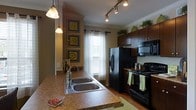 This single-floor space showcases a thoughtfully designed kitchen that balances functionality with aesthetic appeal. The kitchen is anchored by rich, dark wood cabinetry that extends to the upper walls, providing generous storage space. Black appliances, including a French door refrigerator, electric range, and built-in microwave, create a cohesive, modern look throughout the space.

The countertops feature warm-toned granite or granite-style surfaces that complement the dark cabinetry beautifully. A triple-basin stainless steel sink is positioned along the peninsula, which also serves as a functional workspace and casual dining area. Track lighting runs along the ceiling, supplemented by elegant pendant lights that hang over the peninsula, ensuring ample task lighting throughout.

Crown molding adds a refined architectural detail along the ceiling line. Natural light filters through a window dressed with white blinds and sheer curtains, brightening the space. The hardwood flooring grounds the kitchen with warmth, while decorative framed artwork on the adjacent wall adds a personalized touch to the overall ambiance.