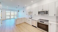 Located at 20 Medford St in Somerville, MA, this thoughtfully designed unit offers four bedrooms and two bathrooms across a single floor. The modern aesthetic is defined by crisp white walls, light hardwood flooring, and clean architectural lines throughout.

The kitchen is a standout feature, boasting white flat-panel cabinetry with matte black hardware, white quartz countertops, and a full suite of stainless steel appliances including a dishwasher, electric range, built-in microwave, and refrigerator. Pendant lighting and recessed fixtures illuminate the space, which opens toward a windowed living area.

The primary bedroom is a comfortable size and benefits from large, floor-to-ceiling windows offering views of the surrounding Somerville neighborhood, flooding the room with natural light. Additional bedrooms are bright and efficiently laid out, with light-filled windows and generous closet space featuring wire shelving systems.

The two bathrooms maintain the home's clean, contemporary palette. One includes a tiled tub-shower combination with matte black fixtures, while the second features a glass-enclosed walk-in shower with white penny tile surround. Both are finished with neutral tile flooring. A connecting hallway provides practical flow between all rooms, completing this well-appointed modern residence.