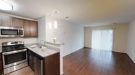 This single-story space showcases an open-concept design that seamlessly connects the kitchen and living area, creating a cohesive and functional layout. The kitchen is outfitted with rich dark wood cabinetry, granite countertops, and stainless steel appliances, including a dishwasher. A peninsula-style counter with a built-in sink serves as a natural divider between the kitchen and the rest of the space, offering additional workspace and a casual spot for seating. Two pendant lights hang above the peninsula, providing focused task lighting while adding a modern decorative touch. The living area extends generously from the kitchen, featuring warm hardwood flooring that runs throughout the entire space, contributing to a consistent and inviting aesthetic. A large sliding glass door fitted with vertical blinds anchors the far wall of the living area, allowing abundant natural light to fill the room while offering access to an outdoor area. The crisp white walls and clean lines throughout maintain a bright, airy atmosphere. A closet near the kitchen adds practical storage to the layout, enhancing the overall functionality of the space.