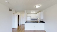 This single-floor space is designed with a functional and clean aesthetic that maximizes usability. The kitchen serves as a central feature, showcasing crisp white cabinetry that extends to the upper walls, providing ample storage. Dark laminate countertops create a striking contrast against the white surfaces, while a peninsula layout offers additional workspace and a natural divider between the kitchen and the adjoining living area. The kitchen is equipped with essential appliances, including a stainless steel refrigerator, a white range, and a built-in microwave, all thoughtfully integrated into the layout. Warm hardwood-style flooring runs throughout the space, adding a sense of warmth and cohesion. The textured ceiling with flush-mount light fixtures ensures even, ambient lighting across the unit. A hallway off the main living area leads to additional rooms and includes a spacious closet with bifold doors, offering generous storage solutions. The open layout, neutral color palette, and practical design elements come together to create a space that is both inviting and highly functional.