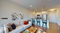 This thoughtfully designed single-floor residence showcases a seamless open-concept layout that connects the living area directly to the kitchen, creating an inviting and functional space. The living room features warm hardwood-style flooring, large windows that welcome natural light, and a sliding door that adds to the sense of openness. A plush sofa, geometric-patterned area rug, and decorative artwork contribute to the space's contemporary aesthetic.

The kitchen is well-equipped with stainless steel appliances, including a refrigerator, electric range, and microwave. Sleek gray cabinetry with brushed nickel hardware is paired with light quartz countertops, while pendant lighting above the breakfast bar adds a refined finishing touch. The bar seating area with black Windsor chairs serves as both a dining and entertaining hub.

The bedroom is a serene retreat, highlighted by a bold charcoal accent wall, plush carpeting, and generous natural light from a large window. A spacious walk-in closet provides ample storage. The bathroom continues the modern design language with gray cabinetry, a wide vanity mirror, quartz countertops, and a glass-enclosed walk-in shower with white tile surround. A stacked in-unit washer and dryer adds everyday convenience to this well-appointed residence.