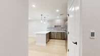 This contemporary single-floor residence is thoughtfully designed with a clean, modern aesthetic throughout. The open-concept kitchen serves as a centerpiece, showcasing two-tone cabinetry with light wood upper cabinets and darker lower cabinets, a striking hexagonal mosaic tile backsplash, crisp white quartz countertops, and a full suite of stainless steel appliances including a refrigerator, range, microwave, and dishwasher. A peninsula with pendant lighting above adds additional prep space and anchors the kitchen to the living area. Light-toned hardwood flooring flows seamlessly throughout the main living areas and bedrooms, lending warmth and continuity to the space. The bedrooms are well-proportioned with neutral gray walls, recessed lighting, and ample room for flexible furniture arrangements. One bedroom features large black-framed floor-to-ceiling windows that frame an expansive urban skyline view, flooding the space with natural light. The bathrooms are finished with large-format dark tile flooring and crisp white walls. The full bath includes a glass-enclosed walk-in shower with classic subway tile surround and a round mirror above a wood-finish vanity. A separate water closet provides added privacy and convenience. White panel doors with matte black hardware add a refined, contemporary touch throughout.