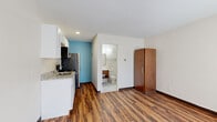 This single-floor studio unit offers a well-organized, open-concept layout that efficiently combines the kitchen and living area into one cohesive space. The rich wood-look vinyl plank flooring runs throughout the main area, adding warmth and visual continuity. Crisp white walls are accented by a bold teal-blue feature wall in the kitchen zone and a matching blue-trimmed entry door, lending the space a modern, distinctive character.

The kitchen is thoughtfully appointed with white shaker-style cabinetry, granite-look countertops, a double-basin stainless steel sink, and a full suite of stainless steel appliances including a refrigerator, range, and over-the-range microwave. A tall wooden wardrobe cabinet provides practical storage in the living area, which also features a wall-mounted PTAC heating and cooling unit beneath a window that allows natural light to filter in.

The full bathroom is finished in clean white tile flooring and features a wood-toned floating vanity with an integrated sink, a framed mirror, and a shower/tub combination with chrome fixtures. The overall design is clean, functional, and modern — making efficient use of every square foot while maintaining a tidy, comfortable aesthetic throughout.