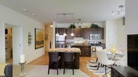 This single-floor residence offers a thoughtfully designed open-concept layout that seamlessly connects the kitchen, dining, and living areas, creating a bright and welcoming atmosphere. The kitchen is a standout feature, showcasing rich dark-toned cabinetry that extends to the upper walls, providing generous storage space, while stainless steel appliances — including a refrigerator, range, and microwave — add a sleek, contemporary touch. A granite-topped kitchen island serves as a central focal point, doubling as a breakfast bar with stylish dark bar seating, ideal for casual dining or entertaining. Pendant lighting hangs gracefully above the island, complementing the track lighting overhead to ensure the space is well-lit throughout. Warm hardwood-style flooring flows across the open area, adding a sense of continuity and warmth to the interior. The dining area is positioned just adjacent to the kitchen, furnished with elegant white upholstered seating and a patterned area rug that adds visual interest. Neutral, light-toned walls enhance the airy feel of the space, while tasteful decorative touches — including potted greenery atop the cabinetry and framed artwork — lend the home a polished, curated character.
