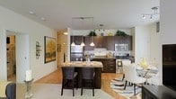 This single-floor residence offers a thoughtfully designed open-concept layout that seamlessly connects the kitchen, dining, and living areas, creating a bright and welcoming atmosphere. The kitchen is a standout feature, showcasing rich dark-toned cabinetry that extends to the upper walls, providing generous storage space, while stainless steel appliances — including a refrigerator, range, and microwave — add a sleek, contemporary touch. A granite-topped kitchen island serves as a central focal point, doubling as a breakfast bar with stylish dark bar seating, ideal for casual dining or entertaining. Pendant lighting hangs gracefully above the island, complementing the track lighting overhead to ensure the space is well-lit throughout. Warm hardwood-style flooring flows across the open area, adding a sense of continuity and warmth to the interior. The dining area is positioned just adjacent to the kitchen, furnished with elegant white upholstered seating and a patterned area rug that adds visual interest. Neutral, light-toned walls enhance the airy feel of the space, while tasteful decorative touches — including potted greenery atop the cabinetry and framed artwork — lend the home a polished, curated character.