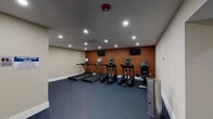 This fitness center is designed to support a comprehensive workout experience within a compact, single-floor layout. The main exercise area is finished with durable speckled blue rubber flooring, providing excellent traction and cushioning underfoot. A bold terracotta accent wall anchors the primary cardio zone, where a row of True-brand treadmills, elliptical trainers, and a stationary bike are neatly arranged side by side. Multiple flat-screen televisions are mounted above the equipment, offering entertainment during workouts. Recessed lighting and a ceiling-mounted HVAC cassette unit ensure consistent illumination and climate control throughout the space. A secondary room houses a multi-function cable strength training machine, a wall-mounted mini-split unit for independent temperature regulation, and a collection of stability and medicine balls for functional training. Natural light filters through a window fitted with horizontal blinds in this area. The facility also includes a dedicated restroom accessible through a connecting corridor, along with a posted fitness center rules sign near the entrance. A stainless steel waste receptacle adds a practical finishing touch. The overall design is clean, functional, and purpose-built to meet a wide range of fitness needs.