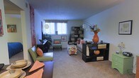 Located at 5125 Cedar Dr in Columbus, Ohio, this single-floor residence offers a comfortable and thoughtfully arranged living space. The living room serves as the heart of the home, featuring wall-to-wall carpeting that adds warmth underfoot and a soft, inviting ambiance throughout. Natural light filters in through a curtained window, brightening the room and highlighting the space's open layout. The living area is generously proportioned, providing ample room for varied furniture arrangements. Décor leans toward an eclectic, earthy aesthetic, with warm tones of orange, green, and brown reflected in the furnishings and accents. Dark-finished shelving units and accent tables provide functional storage and display space, while a bold orange ceramic vase serves as a striking focal point. A mix of seating options, including a sofa, accent chairs, and a floral ottoman, create a welcoming, layered atmosphere. Greenery in the form of indoor plants adds a natural, refreshing element to the interior. The smooth, neutral-toned walls and flat ceiling contribute to a clean, uncluttered backdrop that allows the colorful furnishings to stand out. Overall, the space presents a cozy, well-curated environment suited for comfortable everyday living.