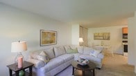 This single-floor residence offers a thoughtfully arranged living space defined by a clean, neutral color palette and an open, airy feel. The living area serves as the heart of the home, centered around a generously sized sectional sofa dressed in soft beige upholstery and accented with decorative throw pillows in blue and green tones. A round dark-wood coffee table anchors the seating arrangement, adding a refined contrast to the light surroundings. Smooth, light-colored walls and ceiling work together to maximize the sense of openness and brightness throughout. Framed artwork adorns the walls, contributing warmth and a curated aesthetic to the space. Transitioning seamlessly from the living area, a dining space is visible in the background, illuminated by a warm table lamp and featuring a clean, minimalist setup. A set of closet or storage doors with warm wood-toned finishes adds both functionality and visual interest. The overall ambiance is calm and inviting, with consistent use of soft tones, tasteful décor, and well-considered furnishings that create a cohesive and comfortable living environment throughout the single-level layout.