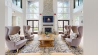 Located at 7000 Johnson Farm Lane in Chadds Ford, PA, this single-floor community space combines classic architectural elegance with thoughtfully modern interior design. The grand entry rotunda sets a welcoming tone, featuring soaring ceilings, white structural columns, light hardwood flooring, and glass double doors that draw the eye toward the main gathering area beyond.

The central lounge is a standout feature, anchored by a floor-to-ceiling stacked stone fireplace flanked by a mounted flat-screen television. Oversized arched windows on either side of the fireplace flood the room with abundant natural light while offering serene views of the surrounding greenery. The seating arrangement — with wingback chairs, plaid accent seating, and a substantial wood coffee table atop a classic patterned area rug — creates an inviting and refined atmosphere.

The kitchenette is equally impressive, finished with white shaker cabinetry, warm butcher block countertops, and gold hardware accents. A textured sage-toned tile backsplash adds a contemporary touch, while open wood floating shelves provide functional display space. A generous window brings in natural light and maintains a bright, airy feel throughout. The space as a whole conveys a warm, upscale ambiance well-suited for community gathering and relaxation.