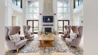 Located at 7000 Johnson Farm Lane in Chadds Ford, PA, this single-floor community space combines classic architectural elegance with thoughtfully modern interior design. The grand entry rotunda sets a welcoming tone, featuring soaring ceilings, white structural columns, light hardwood flooring, and glass double doors that draw the eye toward the main gathering area beyond.

The central lounge is a standout feature, anchored by a floor-to-ceiling stacked stone fireplace flanked by a mounted flat-screen television. Oversized arched windows on either side of the fireplace flood the room with abundant natural light while offering serene views of the surrounding greenery. The seating arrangement — with wingback chairs, plaid accent seating, and a substantial wood coffee table atop a classic patterned area rug — creates an inviting and refined atmosphere.

The kitchenette is equally impressive, finished with white shaker cabinetry, warm butcher block countertops, and gold hardware accents. A textured sage-toned tile backsplash adds a contemporary touch, while open wood floating shelves provide functional display space. A generous window brings in natural light and maintains a bright, airy feel throughout. The space as a whole conveys a warm, upscale ambiance well-suited for community gathering and relaxation.