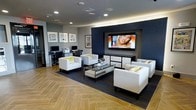 This single-floor space combines a sophisticated lounge area with a functional business center, offering a versatile environment designed for both relaxation and productivity. The lounge is anchored by a bold navy accent wall that frames a large wall-mounted flat-screen television, creating a striking focal point. Crisp white tufted armchairs arranged around a sleek glass-top coffee table provide ample seating, accented with pops of yellow and white throw pillows that add a touch of warmth to the contemporary palette. The flooring features a beautifully laid herringbone wood pattern that transitions seamlessly into a dark area rug beneath the seating arrangement, adding texture and depth to the overall design. Recessed lighting throughout the ceiling ensures the space is well-lit, while subtle cove lighting along the ceiling perimeter adds a warm ambient glow. A collection of framed artwork lines the walls, lending a curated, gallery-like quality to the lounge. Adjacent to the seating area, a dedicated business center offers computer workstations and office equipment, making the space practical for everyday tasks. The clean lines, neutral tones, and modern furnishings come together to create a polished, inviting atmosphere suited for both casual gatherings and focused work.