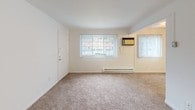 This single-floor residential unit offers a straightforward and functional layout designed for comfortable everyday living. The kitchen is efficiently arranged with warm-toned wood cabinetry, white appliances including a full-size refrigerator and dishwasher, a single-basin sink, and tile flooring — providing ample storage and workspace within a compact footprint.

The living area is bright and open, featuring plush wall-to-wall carpeting, white walls, and multiple windows dressed with vertical blinds that allow natural light to filter through. A baseboard heating unit and a wall-mounted air conditioning unit ensure year-round climate comfort. The entry door opens directly into this welcoming common space.

The bedrooms continue the clean, neutral aesthetic with soft carpeting and crisp white walls. Each room is generously proportioned and includes a built-in closet with shelving and hanging rods, offering practical storage solutions. One bedroom features a window with vertical blinds, bringing in additional natural light.

The bathroom is neatly appointed with a wood-toned vanity, an integrated white sink with standard fixtures, and tile flooring, maintaining the unit's consistent, functional design throughout. A hallway connects the rooms seamlessly, contributing to an efficient and well-organized floor plan overall.