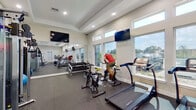 This fitness center is a comprehensively outfitted single-floor space designed to support a full range of workout routines. The room features elevated tray ceilings with recessed lighting, contributing to an open and airy atmosphere throughout. A continuous row of large windows along one wall floods the interior with abundant natural light while offering outdoor views, enhancing the overall workout environment.

The cardio zone is well-represented, with multiple treadmills, elliptical trainers, stair climbers, and stationary cycling bikes arranged to maximize floor space and usability. Multiple wall-mounted flat-screen televisions are positioned throughout the room, providing entertainment options visible from various workout stations.

The strength training area includes a Smith machine, a cable machine system, a dumbbell rack stocked with a full range of free weights, and a weight bench. Wall-to-wall mirrors line one side of the room, visually expanding the space and allowing users to monitor their form during exercises.

Additional fitness accessories, including stability balls, resistance bands, and foam rollers, are stored along the walls. The flooring throughout consists of durable rubber gym matting, providing traction and impact absorption. The clean, neutral color palette of white walls and light trim gives the space a professional and welcoming ambiance.