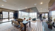 This sophisticated single-floor amenity lounge is designed to impress, combining contemporary architecture with upscale interior design and sweeping views of the Las Vegas Strip. Floor-to-ceiling glass walls and sliding doors flood the space with natural light while framing iconic city landmarks, including the High Roller observation wheel and prominent Strip hotels, creating a seamless connection between the interior and the vibrant cityscape beyond.

The open-plan layout is anchored by a striking geometric-patterned tile floor in neutral tones that transitions elegantly between distinct seating zones. A large communal dining table with upholstered white chairs serves as a central gathering point, complemented by a built-in planter with live greenery. The lounge area features teal velvet sofas, retro-inspired swivel chairs in walnut and black leather, and layered area rugs that define intimate conversation clusters.

Overhead, bold hexagonal pendant light fixtures in warm gold add dramatic visual interest, reinforcing the space's modern aesthetic. High ceilings amplify the sense of openness, while recessed lighting provides ambient illumination throughout. A reception or service counter with teal cabinetry anchors one end of the space, and floor-to-ceiling smoked mirror panels along one wall enhance depth and reflect the panoramic views. A glazed exterior door provides direct access to an outdoor terrace.