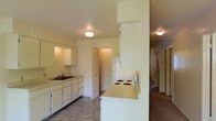 This single-floor space centers around a functional kitchen that serves as the heart of the layout. The kitchen is designed with practicality in mind, featuring white cabinetry along one wall paired with a double sink and countertop workspace. A defining architectural element is the partial dividing wall that incorporates a built-in electric cooktop on its surface, creating a peninsula-style cooking area that separates the kitchen from the adjacent hallway without fully closing off the space.

The flooring in the kitchen area transitions from resilient vinyl-style material to carpet, which continues down the hallway leading to the rest of the unit. The hallway, visible from the kitchen, connects to multiple additional rooms, suggesting a well-organized floor plan. Flush-mount ceiling light fixtures provide consistent overhead lighting throughout, complementing the light-toned walls that give the space a bright, neutral character.

A closet or pantry door is visible near the refrigerator, adding convenient storage. The textured ceiling is a consistent detail throughout. Overall, the space offers a straightforward, functional layout with a connected kitchen and living corridor.
