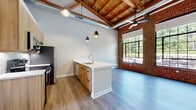 This two-floor loft masterfully blends industrial heritage with contemporary design. The main living area commands attention with its soaring wood beam and plank ceilings, exposed HVAC ductwork, and original brick accent walls that speak to the building's storied past. Large steel-framed windows flood the space with abundant natural light while framing lush outdoor views, and durable light-toned luxury vinyl plank flooring runs throughout both levels.

The open-concept kitchen serves as the heart of the upper floor, anchored by a generous island with a white quartz countertop, an undermount sink, and warm wood cabinetry with matte black hardware. Stainless steel appliances — including a range, microwave, refrigerator, and dishwasher — complete the well-appointed cooking space, which is illuminated by sleek pendant lighting overhead. A staircase connects the floors, leading to additional living areas below.

The bedroom offers a calm retreat, featuring the same exposed wood ceiling and ample floor space. Two bathrooms serve the unit: one with a full tub-shower combination and warm wood-tone flooring, and a second half bath with a modern vanity and frameless mirror. One area of the lower level retains its raw, original concrete and brick walls alongside multi-pane industrial windows, preserving the authentic character of the building's architectural roots.