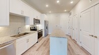 Located at 2400 Linden Dr in Denton, TX, this one-bedroom, one-bathroom unit offers a thoughtfully designed single-floor layout with contemporary finishes throughout. The kitchen serves as a focal point of the home, featuring white shaker-style cabinetry, granite countertops, a subway tile backsplash, a central island, and a full suite of stainless steel appliances including a refrigerator with ice maker, range, microwave, and dishwasher. Light wood-style plank flooring runs continuously throughout the space, creating a cohesive and clean aesthetic. Recessed lighting illuminates the kitchen and main areas effectively. The bedroom is a comfortable, well-proportioned room with a ceiling fan, a window that brings in natural light and outdoor views of surrounding greenery, and direct access to the walk-in closet. The closet offers multiple shelving tiers and hanging rods for organized storage. The bathroom is well-appointed with white cabinetry, granite countertops, a framed mirror, and built-in open shelving for additional storage. An in-unit laundry area adds everyday convenience. The unit also enjoys views of a landscaped outdoor area with trees and green space, visible from the bedroom window and exterior.
