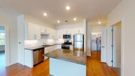 This single-floor residence at The Hills at Port Jefferson offers a well-designed, open-concept layout highlighted by warm hardwood flooring that flows throughout the living areas and bedrooms. The entry corridor, equipped with a built-in intercom panel, leads seamlessly into the main living space, where recessed and track lighting illuminate a clean, neutral interior.

The kitchen is a standout feature, showcasing white shaker-style cabinetry extending to the ceiling, dark granite countertops on both the perimeter surfaces and a central island, and a full suite of stainless steel appliances including a gas range, built-in microwave, refrigerator, and dishwasher. A dedicated utility area off the kitchen houses a stacked washer and dryer. The open living area is generously proportioned, with windows admitting natural light and offering outdoor views of the surrounding greenery.

The bedrooms are well-sized, each featuring hardwood floors, neutral walls, and windows with blinds providing privacy and natural light. The bathroom includes white cabinetry, a granite-topped vanity with an undermount sink, a medicine cabinet with mirrored doors, and a full tub-shower combination. Throughout the residence, the clean white palette, quality finishes, and functional layout create a polished, move-in-ready environment.
