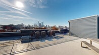 This contemporary rooftop terrace offers a sophisticated urban retreat with breathtaking panoramic views of a bustling city skyline. The space is characterized by its clean lines and modern aesthetic, with light-colored concrete flooring that reflects the abundant natural light. Multiple seating areas are strategically placed to maximize the vista, including a row of sleek, dark-colored lounge chairs perfect for sunbathing or stargazing.

The terrace is partially enclosed by white-painted wooden walls, providing privacy and wind protection while maintaining an open-air feel. A glass door leads to an interior space, seamlessly connecting indoor and outdoor living. The perimeter features a metal railing system that ensures safety without obstructing the spectacular city views.

For entertaining, the space includes a sturdy wooden picnic table, ideal for al fresco dining or casual gatherings. The terrace's layout allows for easy movement and flexible use of the space, whether for solitary relaxation or social events. The unobstructed view of the city's architectural marvels, including prominent skyscrapers and historic buildings, creates a captivating backdrop that changes with the time of day and weather conditions. This rooftop oasis provides a perfect balance of urban living and tranquil escape, offering residents a private perch from which to enjoy the energy of the city below.