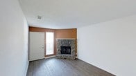 This single-story, 2-bedroom, 1-bathroom home offers a well-organized layout across a comfortable footprint. The living area serves as the heart of the home, highlighted by a striking corner stone fireplace with a white mantel that draws the eye and adds rustic character to the space. Dark hardwood flooring runs throughout, providing a rich, warm contrast against the crisp white walls and a bold terracotta accent wall that adds depth and personality. A textured popcorn ceiling adds a classic finish overhead, while a front-facing window beside the entry door allows natural light to filter into the room. The front door entry is conveniently integrated into the main living space, creating an open and accessible flow from the moment you step inside. The overall ambiance is cozy and inviting, with a blend of natural materials and warm tones that give the home a grounded, lived-in feel. With two bedrooms and a full bathroom rounding out the layout, this home provides practical, comfortable living in a compact and efficiently designed single-floor plan.