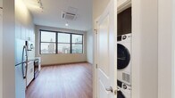 This single-floor unit is positioned at an elevated level, offering sweeping city skyline views through large, dark-framed windows that fill the living spaces with natural light. The open layout features warm hardwood-style flooring that runs throughout the main living areas, creating a cohesive and inviting atmosphere.

The kitchen is thoughtfully designed with white cabinetry extending to the ceiling, light-colored countertops, a subway tile backsplash, and a full suite of stainless steel appliances including a range, microwave, dishwasher, and refrigerator. A dedicated dining area adjacent to the kitchen benefits from expansive windows framing prominent city landmarks.

The bedroom is generously proportioned, with the same hardwood-style flooring and large windows showcasing the urban panorama. A hallway connects the main living spaces to additional rooms, with white panel doors providing access throughout.

The bathroom presents a clean, modern aesthetic with large-format light tile flooring, an elongated white vanity with ample storage, a backlit mirror, and crisp white walls. Conveniently situated nearby is an in-unit stacked washer and dryer, offering added practicality. The overall ambiance is modern, bright, and urban.