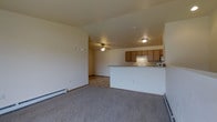 This 2-floor space features an open-concept main living area that seamlessly connects the living room and kitchen. The living room is finished with neutral carpet flooring and warm beige walls, creating a welcoming atmosphere. A ceiling fan is centrally positioned, providing both lighting and air circulation. Baseboard heaters line the walls, offering efficient heating throughout the space.

The kitchen is positioned adjacent to the living area and is defined by a raised breakfast bar that serves as a natural divider between the two spaces. Medium-toned wood cabinetry provides ample storage, while the layout includes essential appliances fitted beneath a range hood. The kitchen benefits from its own ceiling-mounted light fixture, ensuring the workspace is well-lit.

The open sightlines between the kitchen and living area enhance the sense of spaciousness, making the main level feel expansive and connected. The consistent neutral color palette across both areas contributes to a cohesive and clean aesthetic throughout. With two floors, the space offers additional functional areas beyond the main living level.