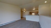 This 2-floor space features an open-concept main living area that seamlessly connects the living room and kitchen. The living room is finished with neutral carpet flooring and warm beige walls, creating a welcoming atmosphere. A ceiling fan is centrally positioned, providing both lighting and air circulation. Baseboard heaters line the walls, offering efficient heating throughout the space.

The kitchen is positioned adjacent to the living area and is defined by a raised breakfast bar that serves as a natural divider between the two spaces. Medium-toned wood cabinetry provides ample storage, while the layout includes essential appliances fitted beneath a range hood. The kitchen benefits from its own ceiling-mounted light fixture, ensuring the workspace is well-lit.

The open sightlines between the kitchen and living area enhance the sense of spaciousness, making the main level feel expansive and connected. The consistent neutral color palette across both areas contributes to a cohesive and clean aesthetic throughout. With two floors, the space offers additional functional areas beyond the main living level.