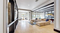 Located at 940 W Canton Ave in Winter Park, Florida, this single-story community clubhouse offers a generously proportioned, open layout designed for both relaxation and social engagement. The interior is anchored by large-format travertine tile flooring that flows seamlessly throughout, reflecting the abundant natural light that pours in through floor-to-ceiling glass doors and expansive windows lining the perimeter.

The space features two distinct living areas. The main lounge is appointed with a large sectional sofa, a plush area rug, and a round wood coffee table, creating an inviting gathering space. Structural columns with dark base accents and exposed dark wood ceiling beams add architectural character and visual depth against the crisp white walls and ceiling.

A standout feature is the dramatic dark-framed built-in media wall, housing a large flat-screen television and open shelving — an elegant focal point that divides the space while maintaining an open, airy feel. A second living area serves as a game room, complete with a full-sized pool table beneath a statement pendant light and a decorative paneled accent wall with an inset display screen.

Glass-paneled doors throughout offer views of the outdoor surroundings, blending the interior with the Florida landscape and filling the space with warmth and light at every turn.
