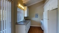 This single-story space showcases a charming and functional kitchen that serves as a central highlight of the home. The kitchen features vaulted ceilings with exposed white-painted wood beams, lending a sense of openness and architectural character to the room. Rich, dark hardwood flooring runs throughout, providing a warm contrast to the crisp white cabinetry that lines the walls. The cabinetry offers generous storage with clean, classic hardware, while the countertops provide ample prep space finished in a neutral stone-toned surface. A double-basin sink is positioned beneath a window fitted with white horizontal blinds, allowing natural light to filter into the space. A row of wall-mounted vanity-style light fixtures above the window provides additional ambient lighting. White appliances, including a range and a full-sized refrigerator, are neatly integrated into the layout, contributing to the kitchen's cohesive and tidy aesthetic. An electrical panel is discretely mounted on the wall, and the sage-green wall color adds a soft, earthy tone that complements the overall warm and inviting atmosphere of the space. The thoughtful design balances practicality with comfort throughout.
