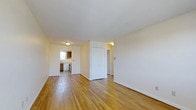 This single-floor space is characterized by its open, airy layout and warm hardwood flooring that runs throughout, lending a cohesive and inviting feel to the interior. The main living area is generously proportioned, with smooth white walls and white baseboards that complement the light-toned wood floors, creating a bright and neutral backdrop suitable for a variety of furnishing styles.

A centrally positioned white door with adjacent wall space divides the layout, providing a natural separation between the living area and the rest of the unit. Beyond this division, a hallway leads further into the space, suggesting additional rooms accessible from the main area.

The kitchen is visible toward the rear, featuring wooden cabinetry in a warm brown tone, a window that allows natural light to filter in, and standard appliances including a stove. The kitchen's layout appears functional and compact, with countertop space along the walls.

Ceiling-mounted light fixtures provide consistent ambient lighting throughout. The overall ambiance of the space is clean, simple, and well-maintained, with a straightforward design that emphasizes practicality and ease of use.