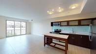This single-floor residence offers a thoughtfully designed open-concept layout with distinct living and kitchen zones unified by light-toned tile flooring throughout the main area. The kitchen is well-appointed with dark walnut cabinetry, frosted-glass upper cabinet doors, a light blue glass tile backsplash, and a full suite of stainless steel appliances including an electric range, built-in microwave, dishwasher, and refrigerator. A freestanding kitchen island with built-in wine storage provides additional workspace and casual dining potential. Track and flush-mount lighting illuminate the kitchen and living area, which opens toward a window allowing natural light to filter in.

The bedroom features plush carpeting, neutral walls, and a generously sized window with an outdoor treetop view, bringing in ample natural light and a connection to the surrounding environment. A hallway off the main living area leads to a full bathroom finished with dark wood vanity cabinetry, a white countertop, tiled shower-tub combination, and tile flooring. A dedicated in-unit laundry closet with a stacked washer and dryer adds everyday convenience. The overall aesthetic is clean, contemporary, and functional.