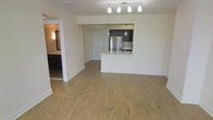This single-floor unit is designed with an open-concept layout that maximizes the sense of space throughout the main living area. Light-toned hardwood flooring extends across the entirety of the space, lending warmth and visual continuity from room to room.

The kitchen is positioned at the rear of the main living area and is defined by a raised white peninsula that serves as both a workspace and a casual dining or seating counter. Dark espresso cabinetry contrasts cleanly against the lighter finishes, while a stainless steel refrigerator adds a modern, polished accent. Pendant lighting hangs above the kitchen zone, complementing a track lighting fixture overhead that illuminates the open living space with directional light.

The front entry door is visible directly from the living area, reinforcing the straightforward, functional layout. To the left, a hallway leads to additional rooms, including what appears to be a bathroom or secondary space with dark cabinetry visible in the background. White walls throughout keep the interior feeling bright and clean, while the neutral palette provides a versatile backdrop suitable for a range of furnishing styles.