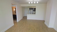 This single-floor unit is designed with an open-concept layout that maximizes the sense of space throughout the main living area. Light-toned hardwood flooring extends across the entirety of the space, lending warmth and visual continuity from room to room.

The kitchen is positioned at the rear of the main living area and is defined by a raised white peninsula that serves as both a workspace and a casual dining or seating counter. Dark espresso cabinetry contrasts cleanly against the lighter finishes, while a stainless steel refrigerator adds a modern, polished accent. Pendant lighting hangs above the kitchen zone, complementing a track lighting fixture overhead that illuminates the open living space with directional light.

The front entry door is visible directly from the living area, reinforcing the straightforward, functional layout. To the left, a hallway leads to additional rooms, including what appears to be a bathroom or secondary space with dark cabinetry visible in the background. White walls throughout keep the interior feeling bright and clean, while the neutral palette provides a versatile backdrop suitable for a range of furnishing styles.