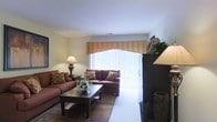 This single-floor space centers around a cozy and well-lit living room that serves as the heart of the home. A large window dressed with an elegant peach valance and vertical blinds allows generous natural light to fill the room, creating a warm and inviting atmosphere. The living area is furnished with plush, earth-toned sofas accented with decorative pillows, arranged around a dark wood coffee table that anchors the space. A classic table lamp adds a soft, ambient glow, complementing the natural light from the window. The neutral-toned walls and light carpeting provide a clean, versatile backdrop that enhances the room's warmth. A dark wood armoire in the corner adds functional storage while contributing to the room's cohesive, traditional aesthetic. Decorative greenery and floral arrangements introduce natural elements, adding texture and life to the interior. The overall ambiance is comfortable and understated, offering a relaxed setting suited for everyday living.