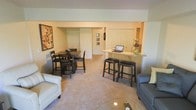 This single-floor space is designed around an open-concept floor plan that integrates the living, dining, and kitchen areas into one cohesive environment. The layout promotes a sense of spaciousness, with neutral-toned carpeting throughout that adds warmth underfoot and ties the zones together visually.

The living area is anchored by a light-colored sofa and a glass-top coffee table, complemented by a second seating option across the room. A large piece of abstract wall art introduces a pop of color against the soft, neutral walls, adding visual interest to the space.

Adjacent to the living area, the dining zone accommodates a dark wood table with matching high-back chairs, suitable for everyday dining or casual gatherings. A peninsula-style breakfast bar with dark upholstered barstools separates the kitchen from the main living space, offering additional seating and a functional workspace. The kitchen features light-toned countertops and built-in storage, maximizing utility within the compact footprint.

An entry corridor with closet storage and framed wall décor leads to the front door, providing a defined transition between the interior and the entrance. Overall, the space presents a clean, functional, and cohesive design.