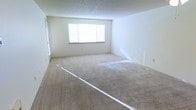 This single-floor space presents a generously sized open living area finished with wall-to-wall beige carpeting that adds a warm, neutral foundation throughout. The room is characterized by its clean, minimalist aesthetic, with crisp white walls that enhance the sense of openness and brightness. A centrally positioned window allows natural light to filter into the space, casting soft illumination across the carpet and contributing to an airy atmosphere. The textured ceiling adds a subtle layer of dimension to the room's overall finish. The layout is straightforward and versatile, offering ample room for a variety of furniture arrangements to suit different lifestyle needs. A doorway visible along one wall suggests connectivity to additional areas of the unit, allowing for natural flow between spaces. The overall condition of the space appears well-maintained and move-in ready, with no significant wear visible on the walls or flooring. The neutral color palette throughout makes the space highly adaptable to a range of interior design preferences, providing a blank canvas for personalization.