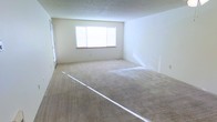 This single-floor space presents a generously sized open living area finished with wall-to-wall beige carpeting that adds a warm, neutral foundation throughout. The room is characterized by its clean, minimalist aesthetic, with crisp white walls that enhance the sense of openness and brightness. A centrally positioned window allows natural light to filter into the space, casting soft illumination across the carpet and contributing to an airy atmosphere. The textured ceiling adds a subtle layer of dimension to the room's overall finish. The layout is straightforward and versatile, offering ample room for a variety of furniture arrangements to suit different lifestyle needs. A doorway visible along one wall suggests connectivity to additional areas of the unit, allowing for natural flow between spaces. The overall condition of the space appears well-maintained and move-in ready, with no significant wear visible on the walls or flooring. The neutral color palette throughout makes the space highly adaptable to a range of interior design preferences, providing a blank canvas for personalization.