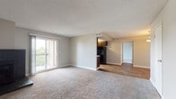 This single-floor unit offers a well-organized layout with distinct living, kitchen, and private spaces. The spacious living room is finished with neutral-toned carpet and features a black-faced fireplace along one wall, adding a functional focal point to the room. A sliding glass door floods the space with natural light and opens to a private balcony with outdoor views of surrounding greenery. A textured ceiling runs throughout the main living area, contributing to the room's understated character.

The galley-style kitchen is positioned just off the living area, featuring dark cabinetry on both sides, white countertops, and black appliances including a refrigerator and dishwasher. A window above the counter provides natural light and an exterior view. The kitchen transitions seamlessly to the rest of the unit via wood-look flooring, which continues into the hallway and secondary spaces.

The bathroom is clean and functional, equipped with a full tub and tiled shower surround, a toilet, chrome fixtures, and a vanity with a light countertop. Wood-look flooring extends through this space as well. An additional area with two doors—likely closet and entry access—is illuminated by a pendant light, completing the unit's practical and cohesive interior design.