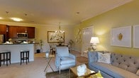 This single-floor residence presents an inviting open-concept layout that seamlessly connects the living, dining, and kitchen areas into one cohesive living space. The warm yellow walls create a bright, cheerful ambiance throughout, complemented by crisp white crown molding that adds a touch of classic architectural elegance.

The living area is tastefully appointed with a tufted gray sofa and a coordinating armchair, arranged around a mirrored gold coffee table. A patterned area rug anchors the seating arrangement, while a table lamp provides soft, ambient lighting. Decorative wall accents add a stylish focal point beside the entry door.

The adjacent dining area features a dark-toned table with upholstered chairs beneath a classic multi-arm chandelier, creating an intimate setting for meals. Framed artwork and potted plants enhance the refined yet welcoming atmosphere.

A glimpse of the kitchen reveals dark cabinetry and a counter bar area. Neutral-toned carpeting flows throughout the main living spaces, contributing to the overall warmth of the home. The space balances comfort and style with its cohesive color palette and well-considered furnishings.