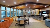This single-floor amenity space is designed to serve as a social hub, combining comfort and entertainment in a cohesive, well-appointed layout. The room is anchored by large floor-to-ceiling windows along one wall, flooding the interior with natural light and offering views of the outdoors, creating an open and airy atmosphere even during evening hours.

The space is organized into several distinct functional zones. A billiards table takes center stage in the main gathering area, surrounded by ample room for play and socializing. Nearby, multiple dark-wood dining tables with upholstered chairs provide comfortable seating for groups of varying sizes. A wall-mounted flat-screen television is positioned to be visible from much of the room, making it ideal for communal viewing.

Along one side of the space, a sleek bar counter with a row of tall bar stools offers an additional seating option, complemented by pendant lighting that adds a warm, inviting glow. Recessed ceiling lights and decorative fixtures are layered throughout, creating a dynamic and welcoming ambiance. The ceiling features bold architectural beams with contrasting tones, adding depth and character to the overall design.

The flooring transitions between carpeted and hard surface areas, subtly defining each zone while maintaining a unified aesthetic throughout this versatile, entertainment-focused space.